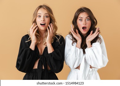 Portrait of two shocked women in black and white clothes looking at camera with throwing up hands isolated over beige background