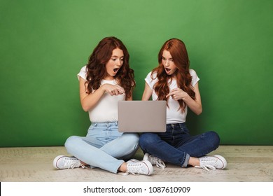 Portrait of two shocked young redhead girls pointing finger at laptop computer while sitting on a floor isolated over green background - Powered by Shutterstock