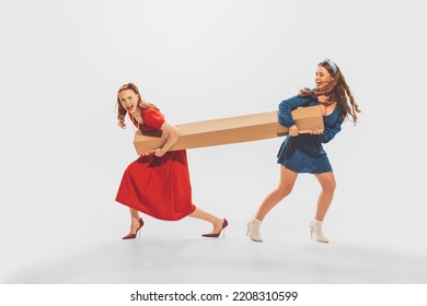 Portrait Of Two Stylish Beautiful Girls Pulling On Big Cardboard Box Isolated Over White Background. Big Sales Hunting. Black Friday Shopping . Concept Of Retro Fashion, Youth Culture, Emotions, Ad