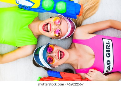 Portrait Of Two Trendy Cool Hipster Girls In Bright Lime And Pink Swimsuit Holding Colorful Water Pistols, Guns, Trendy Hairstyles And Makeup, Sunglasses, Laughing, Summer, Crazy Emotions, Top View