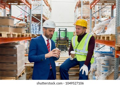 Portrait Of Two Warehouse Workers Taking Break Sitting On Pellets Drinking Coffee And Chatting