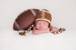 Portrait Of A Two Week Old, Sleeping Newborn Baby Boy. He Is Posed With An American Football Wearing A Crocheted Football Hat.