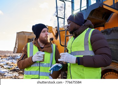 Portrait Of Two Workers, One African-American, Drinking Coffee And Chatting Next To Heavy Industrial Truck On Worksite