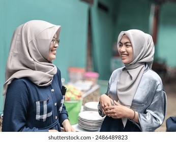 Portrait of two young Asian women in hijabs chatting cheerfully, standing and laughing, against a blurred background - Powered by Shutterstock