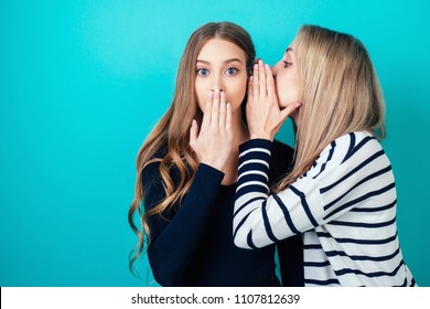 portrait of two young attractive happy girlfriends women with makeup eavesdrops whispers a secret (mystery) in the studio on a blue background. the concept of gossip and confidentiality
