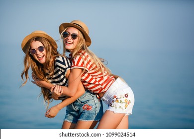 Portrait of two young beautiful girls in trendy stripe shirt, beach hat, sunglasses an jeans shirt on the sea. two friends having fun at the sea with cocktails. Youth and happiness concept.