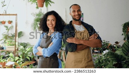 Portrait of two young beautiful professional florist in aprons standing at own small flower shop and smiling to camera. African american couple posing in front of camera concept of family business and