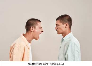 Portrait Of Two Young Caucasian Men, Twin Brothers Arguing, Shouting While Standing Face To Face Isolated Over Beige Background. Family Relationships Concept