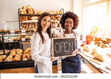 Portrait of two young entrepreneurs standing in the bakery shop with OPEN sign. Two cheerful small business owners smiling and looking at camera while standing with open sign board. - Powered by Shutterstock