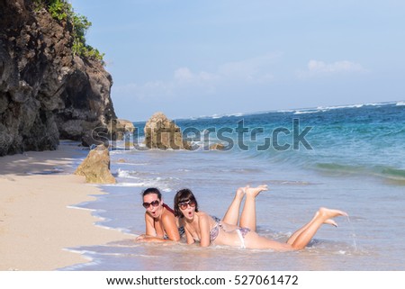 Portrait of two young female friends lie on the sea shore looking at camera and laughing. Caucasian young women strolling along a beach. Tropical island Bali, Indonesia.