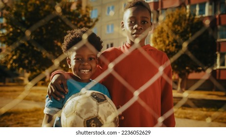 Portrait of Two Young Handsome Black Boys Embracing Each Other while Looking at Camera and Smiling. Younger Brother Holding a Soccer Ball. African Kids Standing in Urban Backyard Together. - Powered by Shutterstock