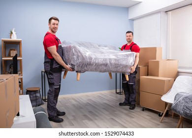 Portrait Of Two Young Happy Male Movers Carrying Wrapped Sofa In New House