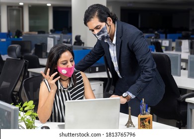 Portrait Of Two Young Indian Businesspeople Wearing Covid Protection Mask And Discussing Project At Workstation, Corporate Environment.