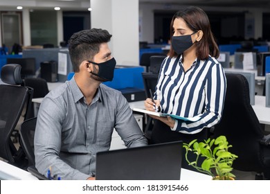 Portrait Of Two Young Indian Businesspeople Wearing Covid Protection Masks While Discussing Project At Workstation, Corporate Environment.