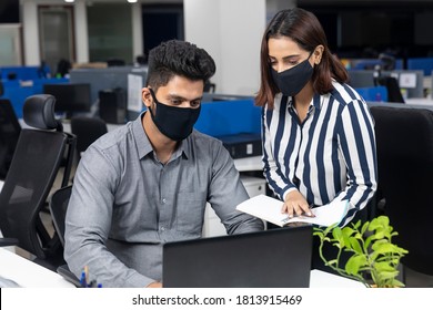 Portrait Of Two Young Indian Businesspeople Wearing Covid Protection Masks While Discussing Project At Workstation, Corporate Environment.