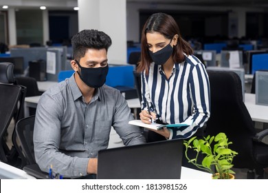 Portrait Of Two Young Indian Businesspeople Wearing Covid Protection Masks While Discussing Project At Workstation, Corporate Environment.