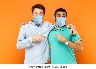 Portrait Of Two Young Worker Men With Surgical Medical Mask Standing, Hugging, Showing And Pointing Each Other And Looking At Camera With Funny Face. Indoor Studio Shot Isolated On Orange Background.