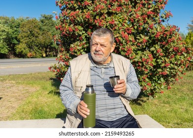 Portrait of ukrainian senior man sitting on  bench against rowan tree and holding vacuum bottle getting ready to drink tea at sunny autumnal day - Shutterstock ID 2159493265