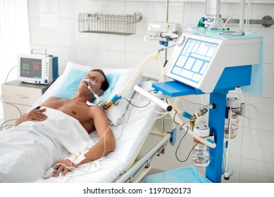 Portrait Of Unconscious Middle Aged Man On Mechanical Ventilator Lying In Hospital Bed Under White Sheets