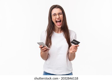 Portrait of uneasy annoyed and angry young brunette female in t-shirt yelling pissed-off as checking bank account, have no money on credit card, or it expired, hold mobile phone, white background