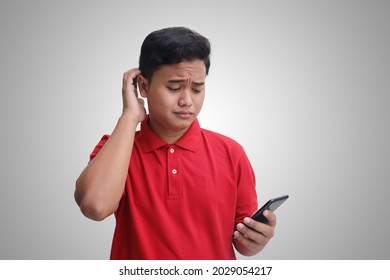 Portrait Of Unhappy Asian Man In Red Polo Shirt Holding Mobile Phone With Sad Expression On Face While Scratching Forehead. Isolated Image On Gray Background