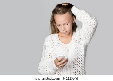 Portrait of unhappy beautiful casual young lady wearing white knitted sweater, looking at smartphone with troubled expression, reading message, waiting for call, studio, gray background, copy space