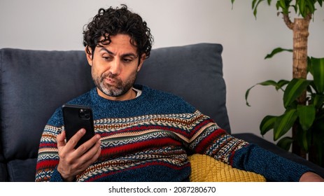 Portrait Of Unhappy Caucasian Man Using Smartphone With Embarrassing Expression. Serious Man Checks Phone Device With Surprise Expression On Couch At Home Interior. Negative Disguting Emotion Concept