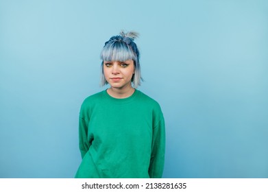 Portrait of unhappy girl in green sweatshirt and blue hair standing and looking at camera with serious face isolated on blue background. - Powered by Shutterstock