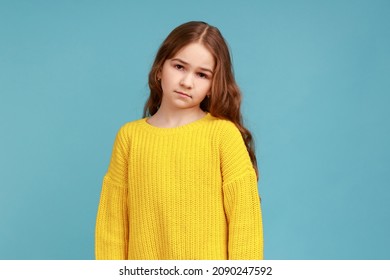 Portrait of unhappy little girl looking at camera with sad upset expression, being in bad mood, wearing yellow casual style sweater. Indoor studio shot isolated on blue background.