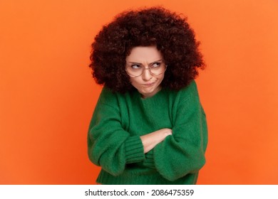 Portrait of unhappy offended woman with Afro hairstyle in green casual style sweater standing with folded hands, looking away, having sad expression. Indoor studio shot isolated on orange background.
