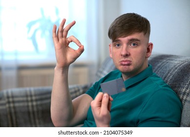 Portrait Of Unhappy Poor Sad Frustrated Upset Guy, Young Handsome Man Who Lost All His Money Holding Bank Credit Card Sitting On Couch At Home And Show Zero Sign. Internet Fraud, Unemployment Concept.