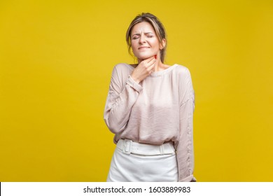 Portrait Of Unhappy Sick Woman With Fair Hair In Casual Beige Blouse Standing With Closed Eyes, Touching Neck Suffering Sore Throat, Flu Symptoms. Indoor Studio Shot Isolated On Yellow Background