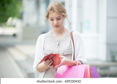 Portrait Of Unhappy Stressed Beautiful Person Looking In Open Wallet With Shocked Expression While Holding Color Shopping Bags At Mall Entrance. Young Model Spent Too Much Money During Shopping Time