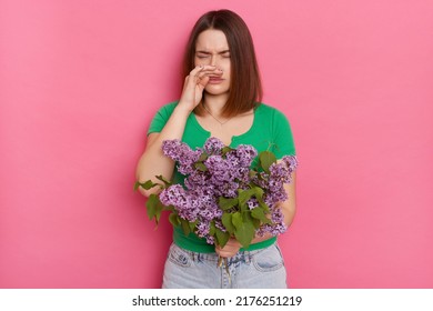 Portrait Of Unhappy Woman Wearing Green T-shirt Posing Isolated Over Pink Background, Standing And Holding Bouquet Of Lilac Flowers, Touching Nose, Having Allergy Symptoms.