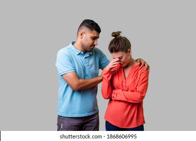 Portrait Of Unhappy Worried Young Couple In Casual Wear Standing Together, Man Comforting Crying Woman, Holding Her Shoulders, Expressing Sympathies. Isolated On Gray Background, Indoor Studio Shot