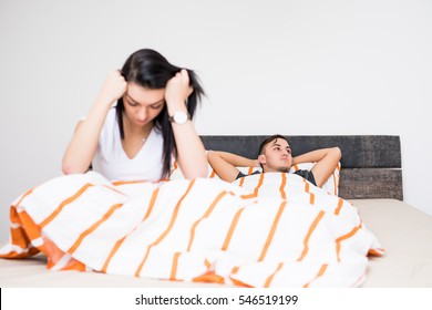 Portrait Of Unhappy Young Heterosexual Couple In Bedroom. Sad Woman In Focus, Dissapointed Man On Back.