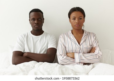 Portrait Of Unhappy Young Mixed Race Couple Posing In Bedroom. Angry Black Guy Sitting On Bed Next To His Mad Wife Keeping Arms Folded, Ignoring And Not Talking To Each Other, Having Disagreement