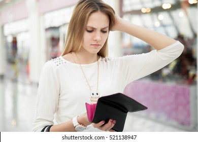 Portrait of unhappy young woman looking in her wallet in shopping center, spent too much, not enough cash, lost money, broke. Shopping center with shops on the background