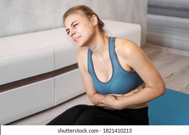 Portrait Of Unhappy Young Woman Sitting On Yoga Mat, Touching Her Stomach After Training, Suffering From Stomach, Feeling Pain, Side View