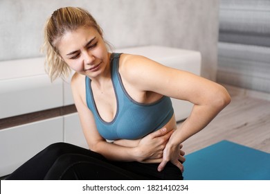 Portrait Of Unhappy Young Woman Sitting On Yoga Mat, Touching Her Back After Training, Suffering From Backache, Feeling Pain, Side View