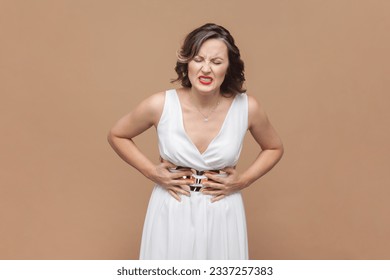 Portrait of unhealthy sick middle aged woman with wavy hair standing, holding her belly with hands, stomach cramps, wearing white dress. Indoor studio shot isolated on light brown background. - Powered by Shutterstock