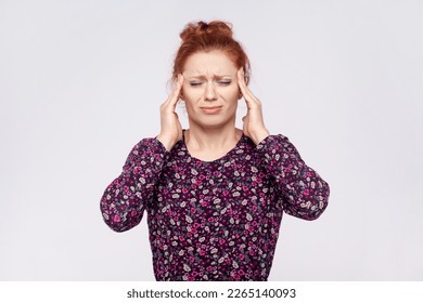 Portrait of unhealthy sick young adult redhead woman wearing dress massaging temples, suffering from headache, migraine, feels unwell. Indoor studio shot isolated on gray background. - Shutterstock ID 2265140093