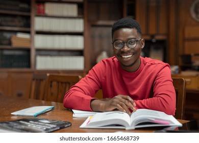 Portrait Of University Student Doing Homework In School Library And Smiling. Happy High School Student Looking At Camera While Studying For Exam. African American Guy With Open Book And Copy Space.