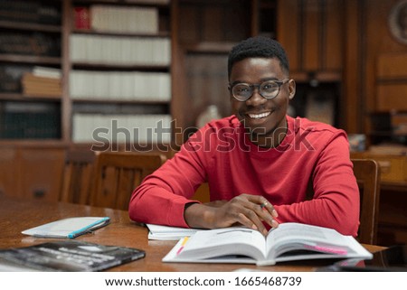 Portrait of university student doing homework in school library and smiling. Happy high school student looking at camera while studying for exam. African american guy with open book and copy space.