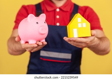 Portrait Of Unknown Anonymous Man Wearing Blue Uniform And Red T-shirt Showing Piggy Bank And Paper House, Saving Money Buying Real Estate. Indoor Studio Shot Isolated On Yellow Background.