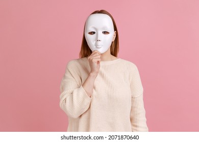 Retrato de una mujer anónima desconocida cubriendo su cara con máscara blanca, escondiendo su verdadera personalidad, anonimato, usando suéter blanco. Estudio cubierto aislado en un fondo rosado.