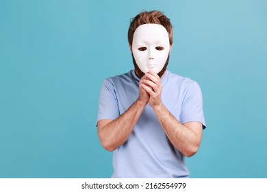 Portrait of unknown man wearing T-shirt covering his face with white mask, hiding his real mystery personality, fake, anonymity. Indoor studio shot isolated on blue background.