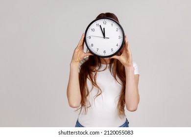 Portrait Of Unknown Woman Holding Wall Clock, Hiding Her Face, Time Management, Schedule And Meeting Appointment, Wearing White T-shirt. Indoor Studio Shot Isolated On Gray Background.