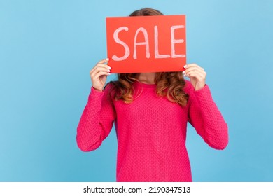 Portrait of unknown woman wearing pink pullover, covering face with Sale inscription, market discounts, black friday, low prices. Indoor her studio shot isolated on blue background.