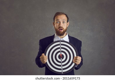 Portrait of unlucky young businessman caught in dangerous trap set up by rivals. Scared man holding dartboard goal becomes target as metaphor of risky business, threat and bad situations at work